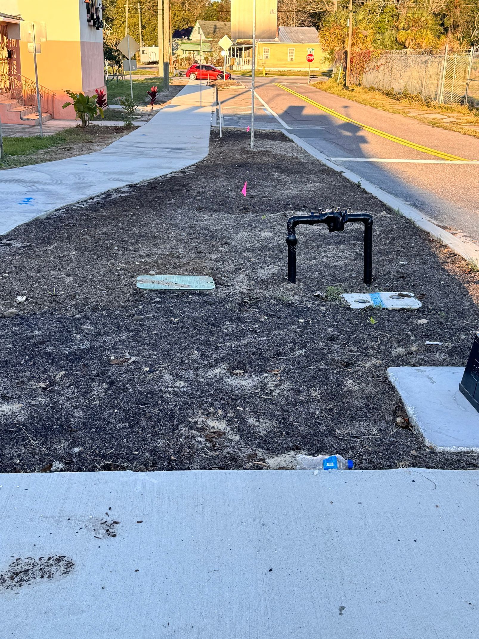 Gravelly ground with utility structures beside a sidewalk and road. Beige buildings in the background.