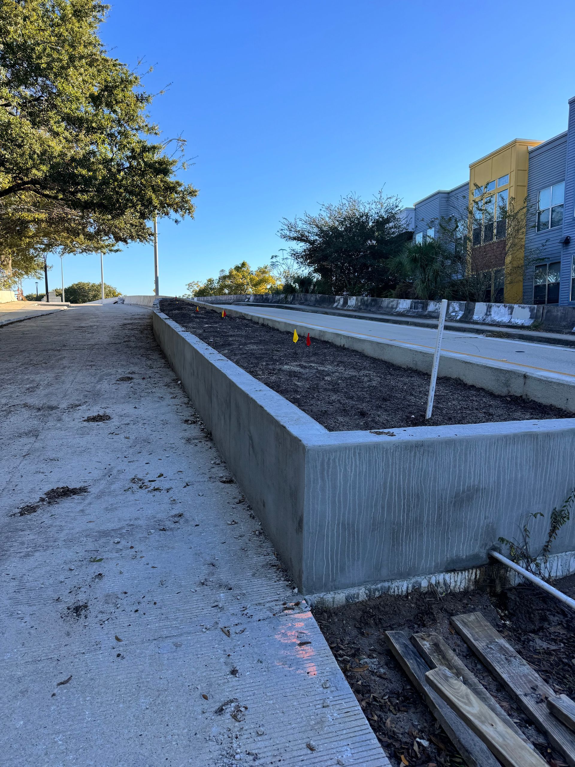 Concrete raised garden beds filled with soil. A paved pathway and modern building in the background. Sunny day.