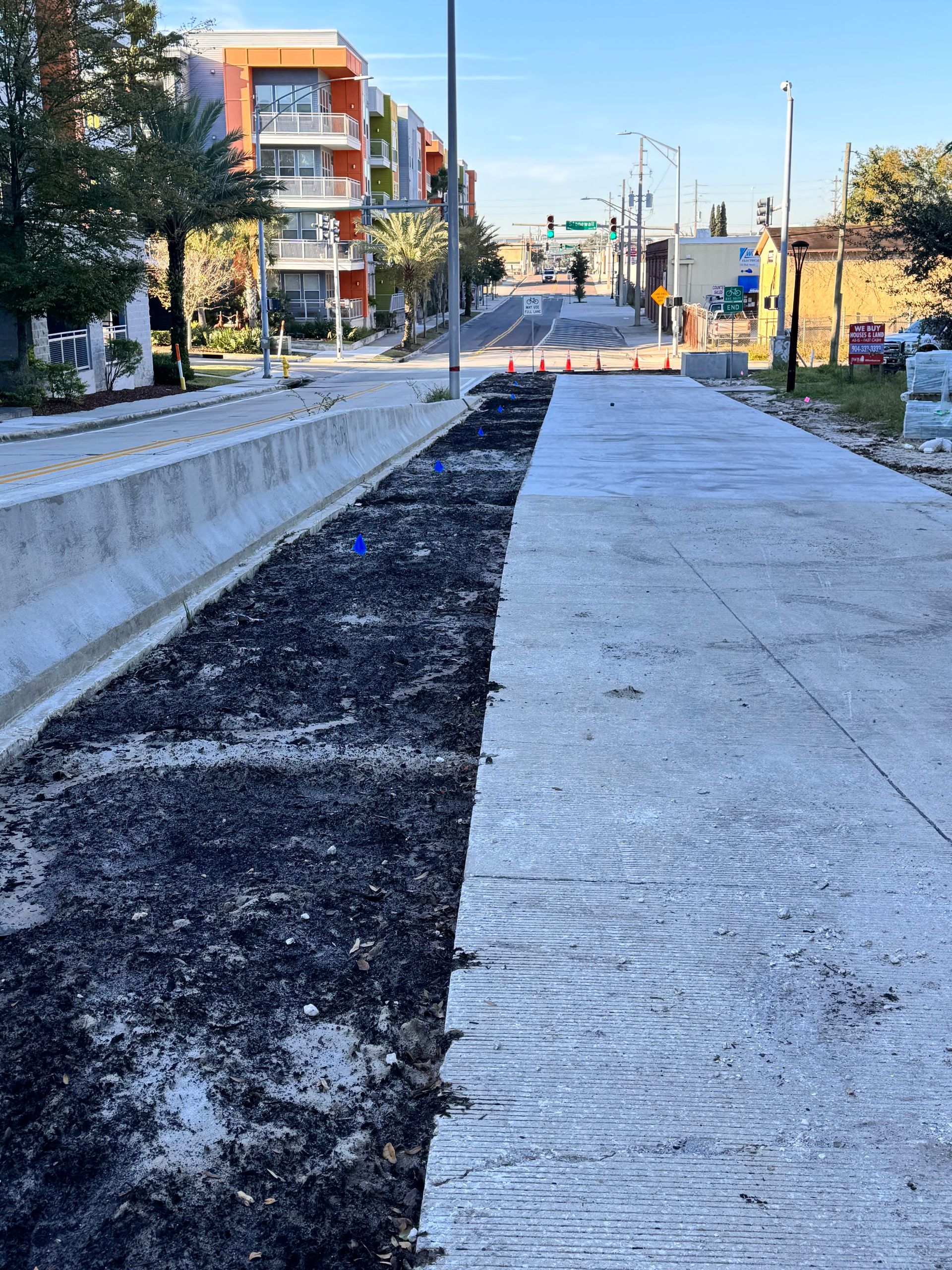 Street construction scene: unfinished road with dirt, concrete barriers, and buildings.