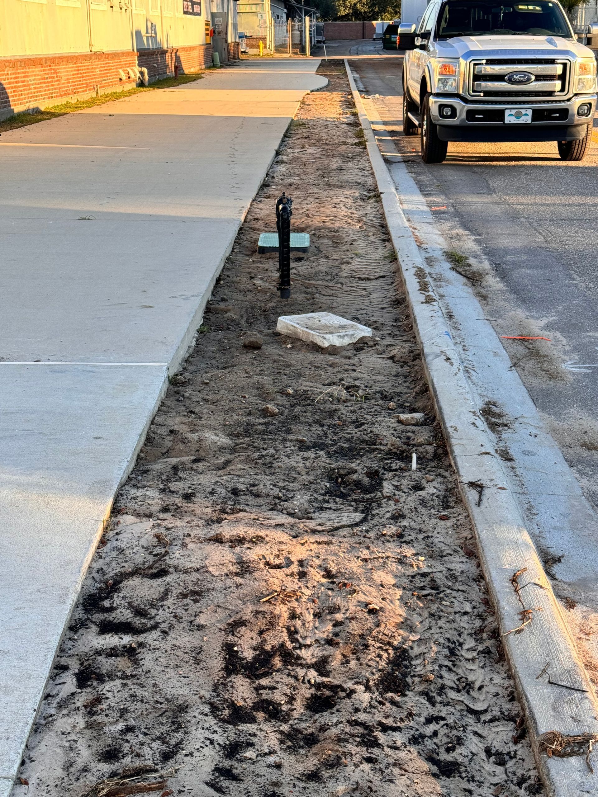 Construction of a curb-side planting area. Dirt trench between a sidewalk and street with a utility post and truck parked nearby.