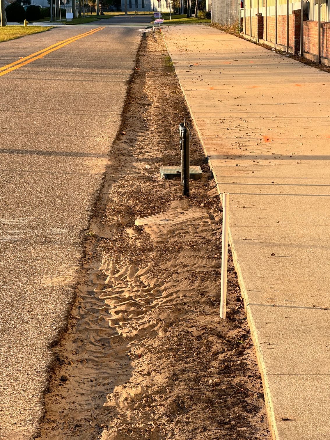 Sidewalk trench with exposed dirt and utility pipes beside a paved street, next to a brick wall.