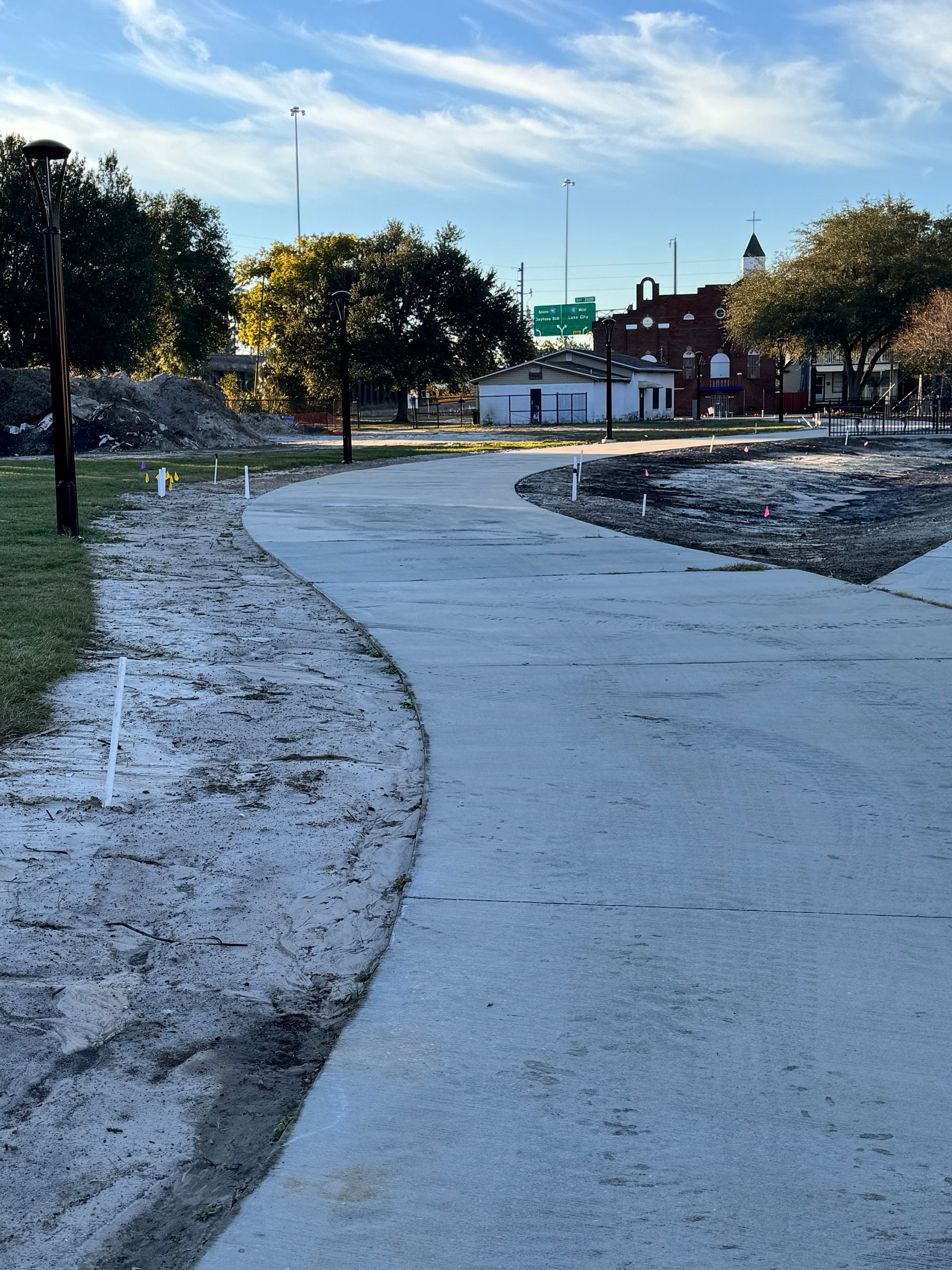 Concrete pathway curves through a park with scattered construction debris, trees, and buildings in the background.