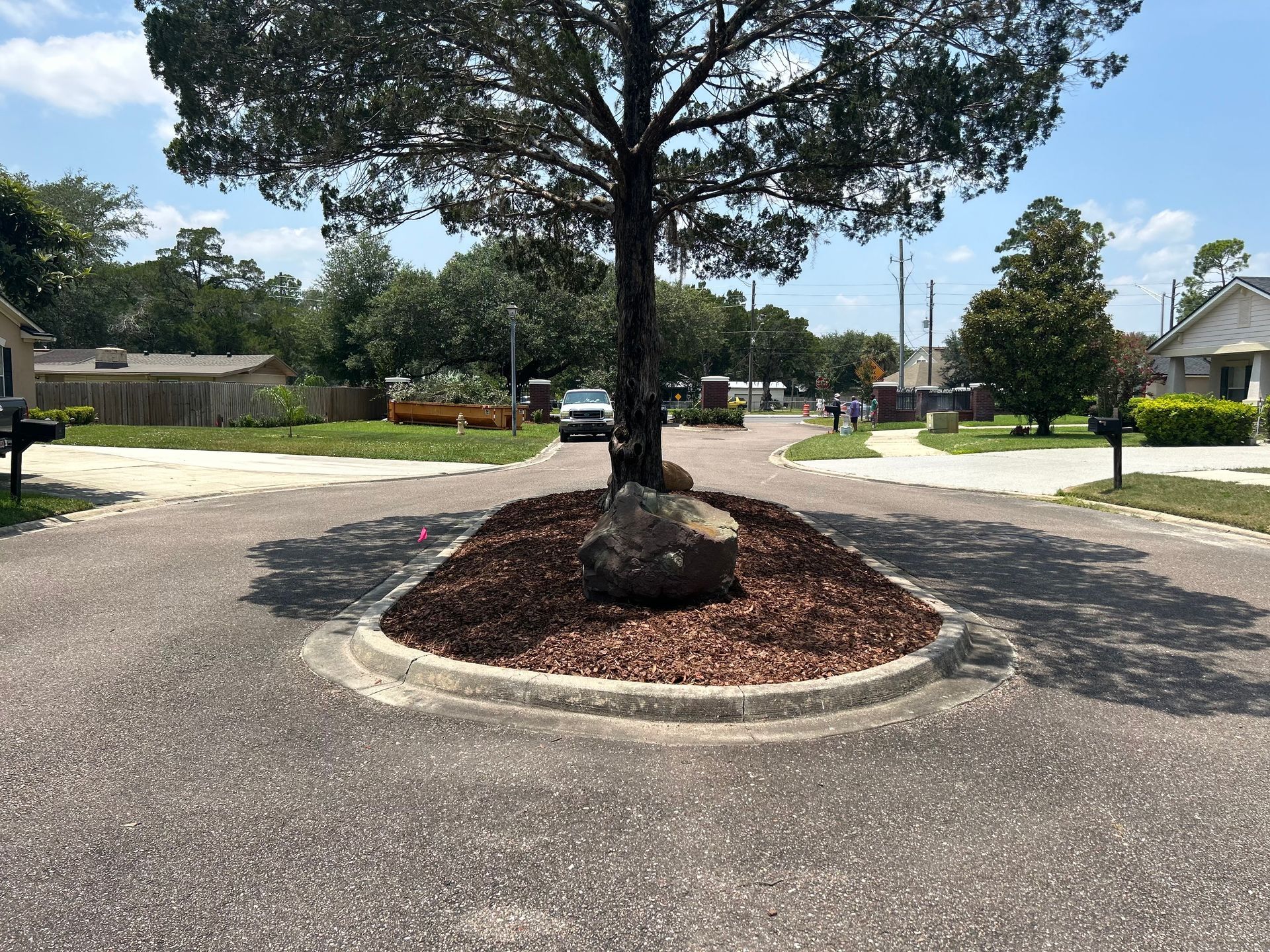 A tree with a large rock in a roundabout in a residential area.