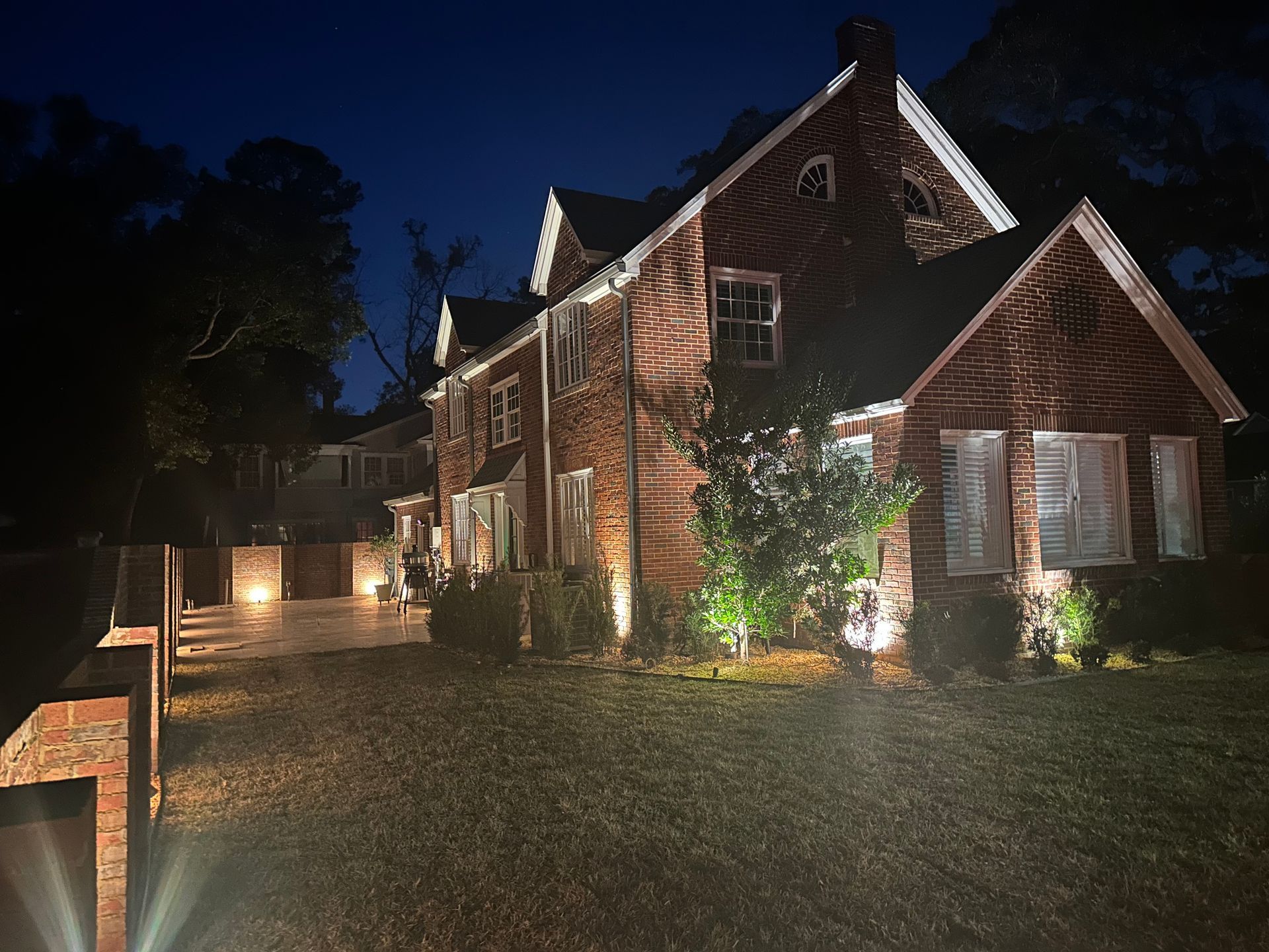 Nighttime view of a brick house with lit windows, a driveway, and glowing landscape lights in front