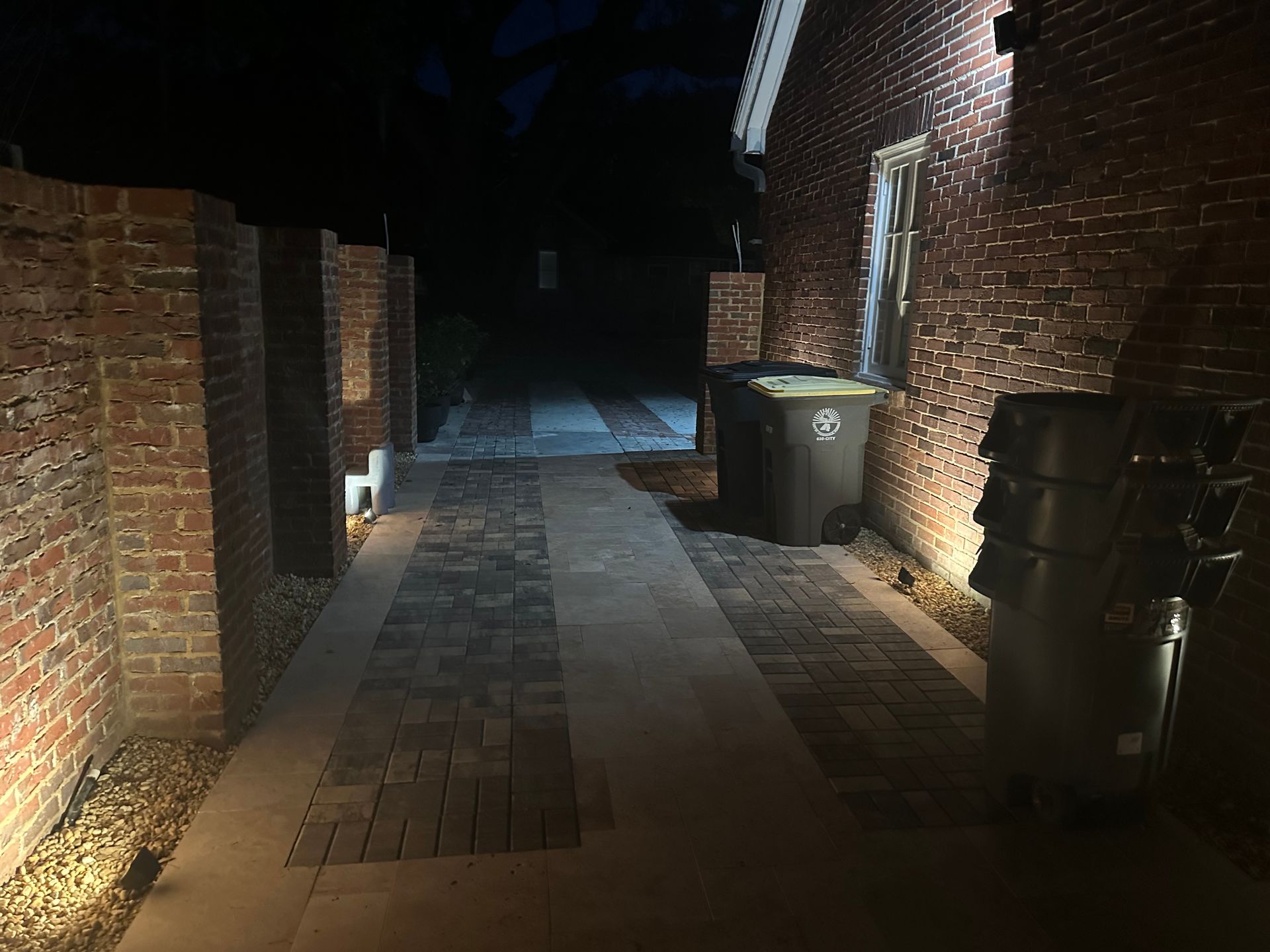 Brick walkway at night, lit by spotlights.  Brick walls flank the path with a dark alleyway ahead.