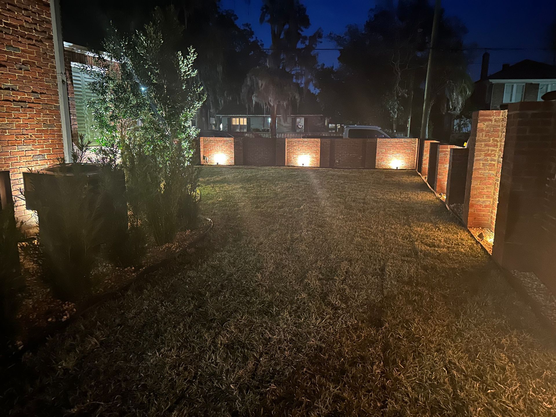 Exterior view at night of a brick wall with illuminated square lights. Grass and bushes are in the foreground.
