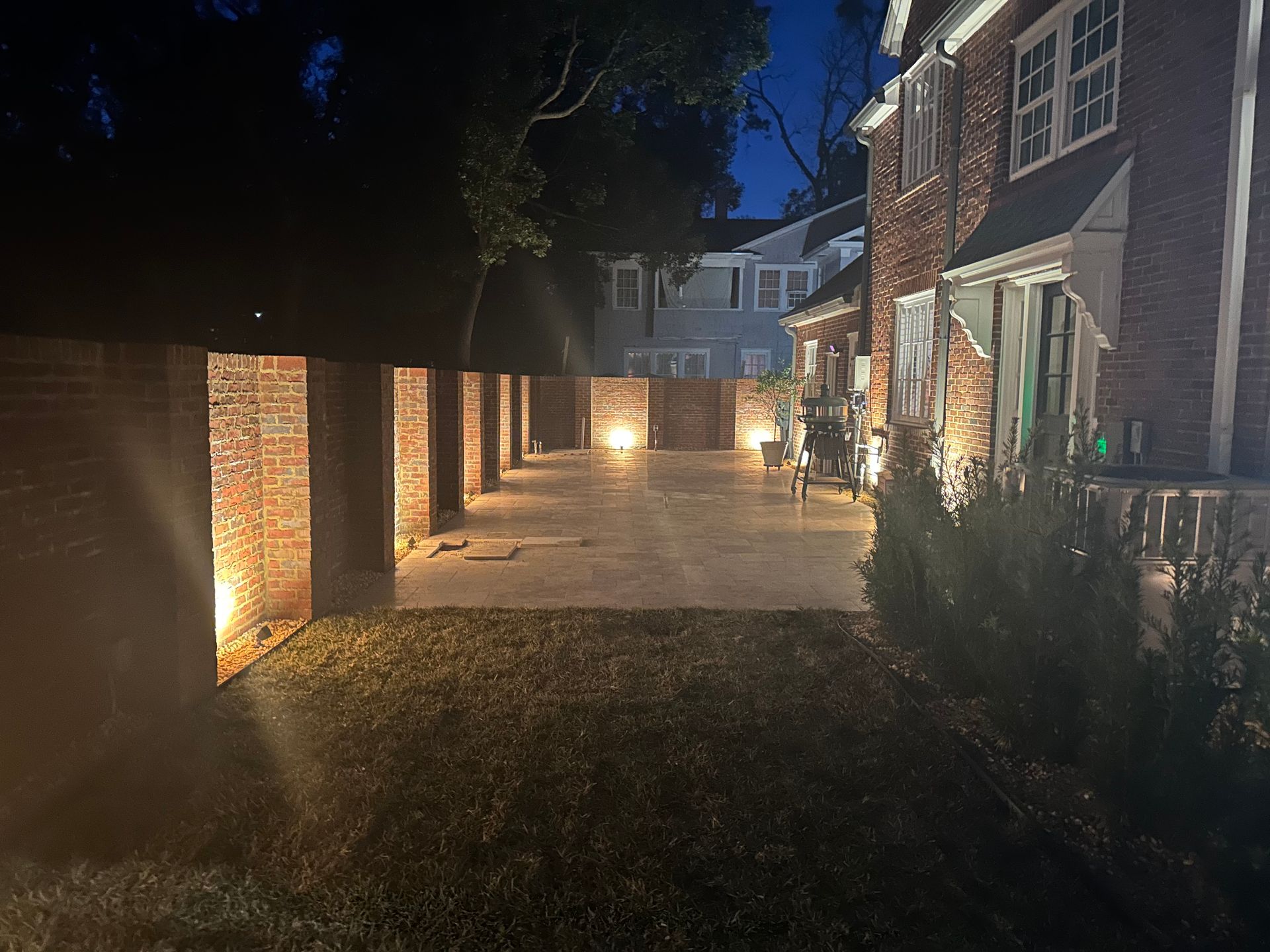 Exterior night view: brick patio area lit by spotlights, adjacent to a brick house. Trees in the background.