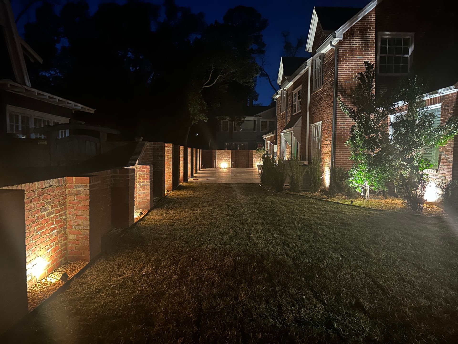 Night view of a brick house and pillars illuminated by ground lights. A grassy lawn leads to a paved patio.