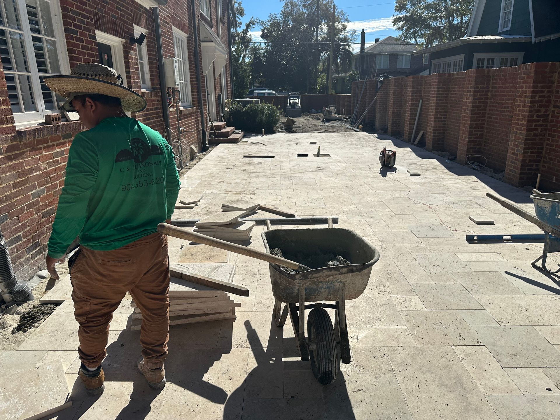 Construction worker near a brick building and a wheelbarrow filled with mortar, laying paving stones.