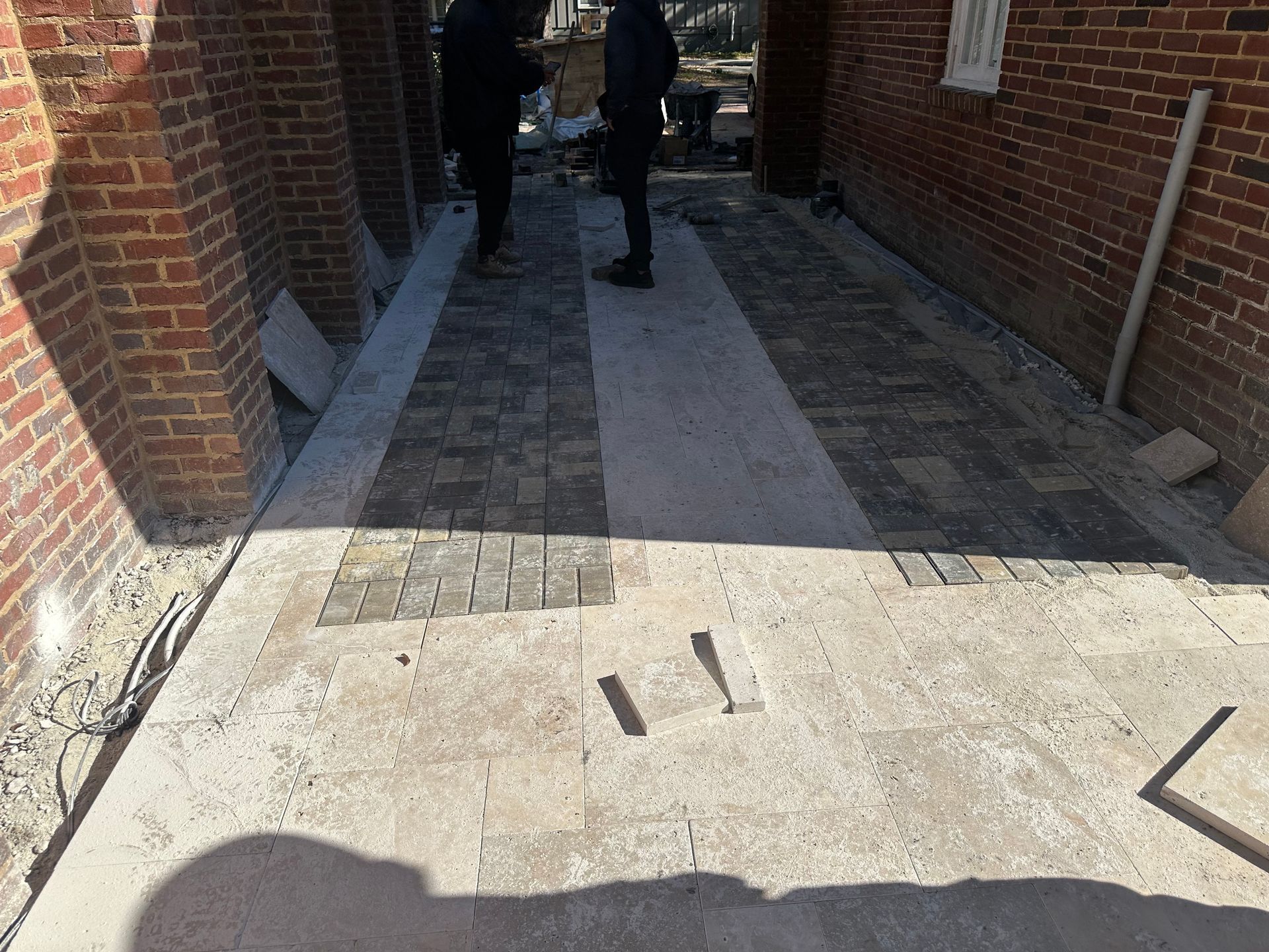 Driveway under construction with brick walls, two people standing, unfinished paving.