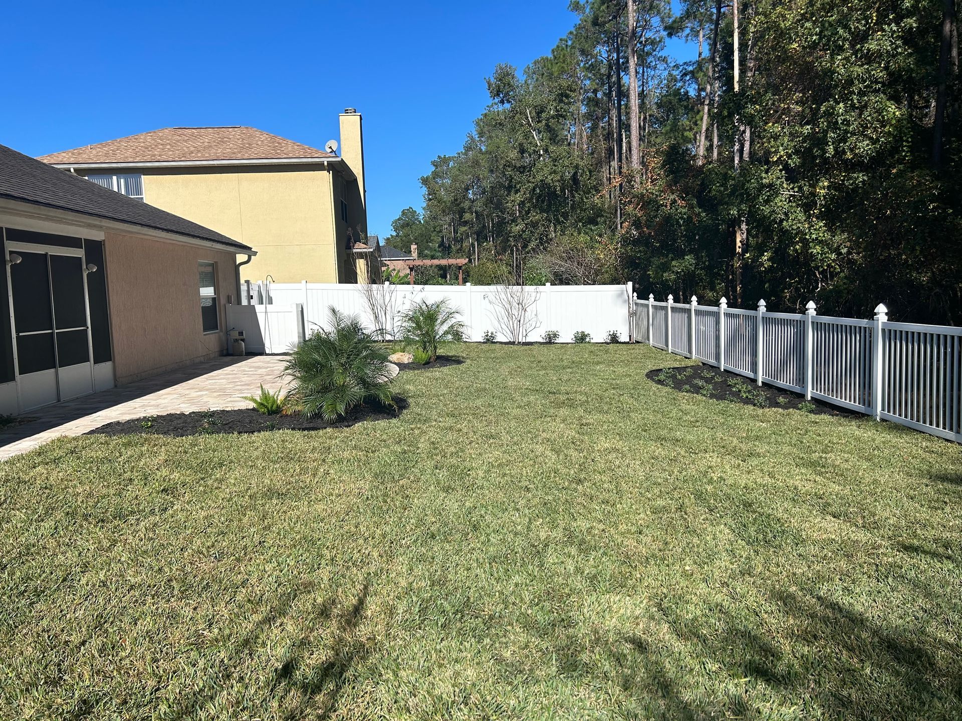 Lawn with white fence, palm trees, and beige house on a sunny day.
