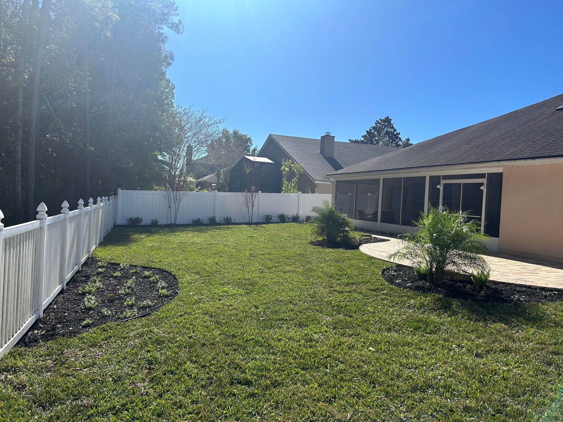 Backyard with a white fence, green grass, and a screened-in porch. Sunny day.