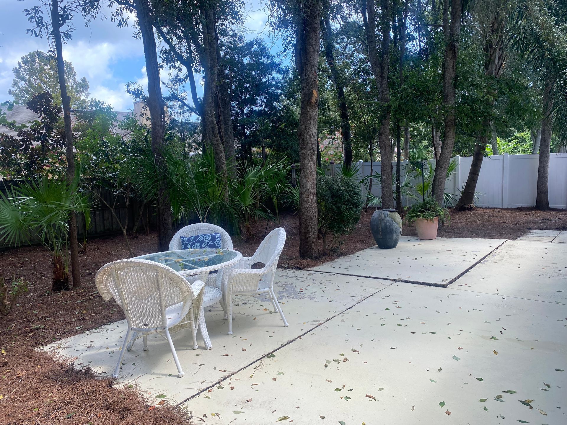 Patio with white table and chairs, surrounded by trees and greenery, next to a white fence.