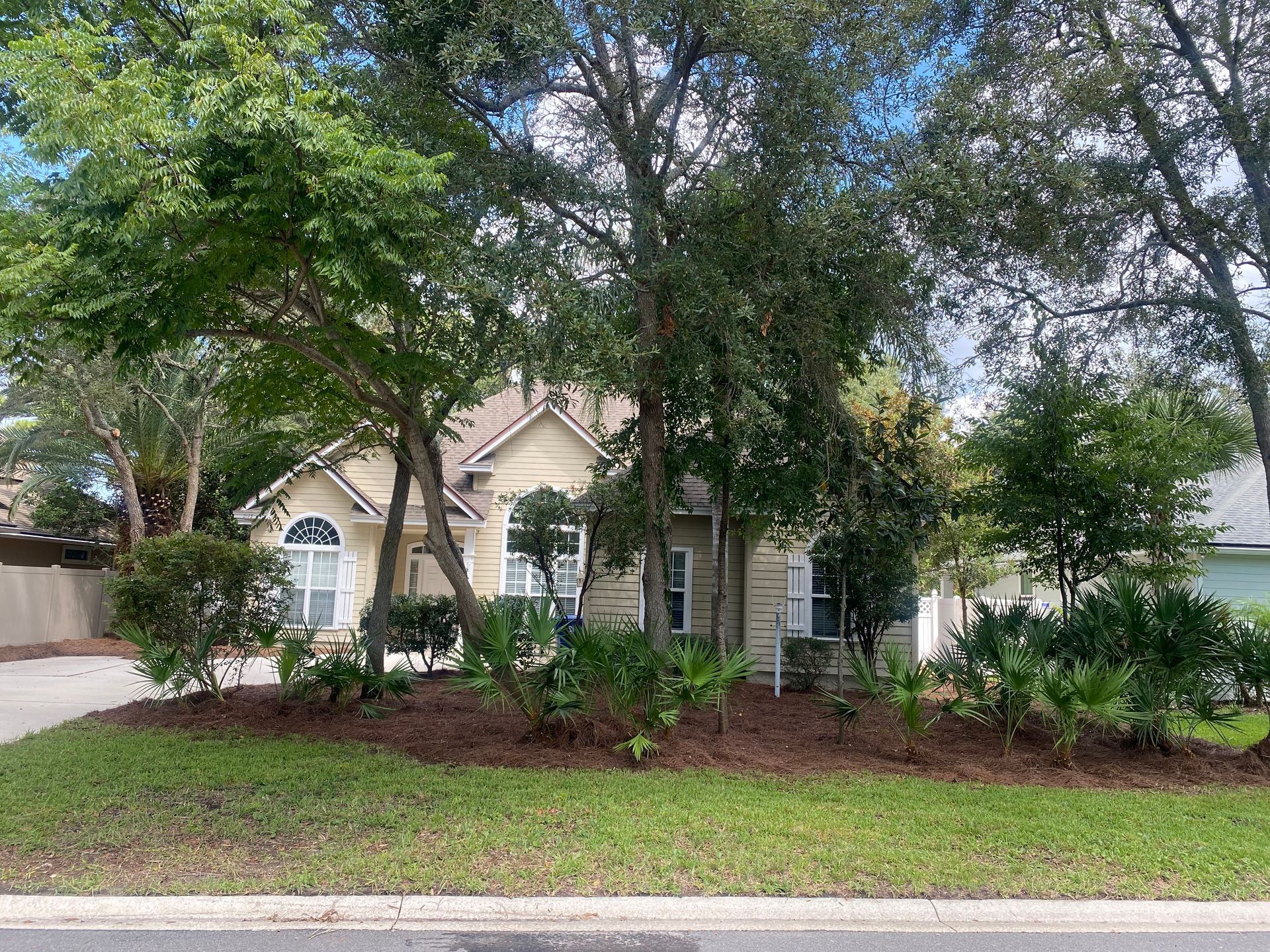 House with beige siding, framed by green trees and landscaping with brown mulch; on a grassy lawn.