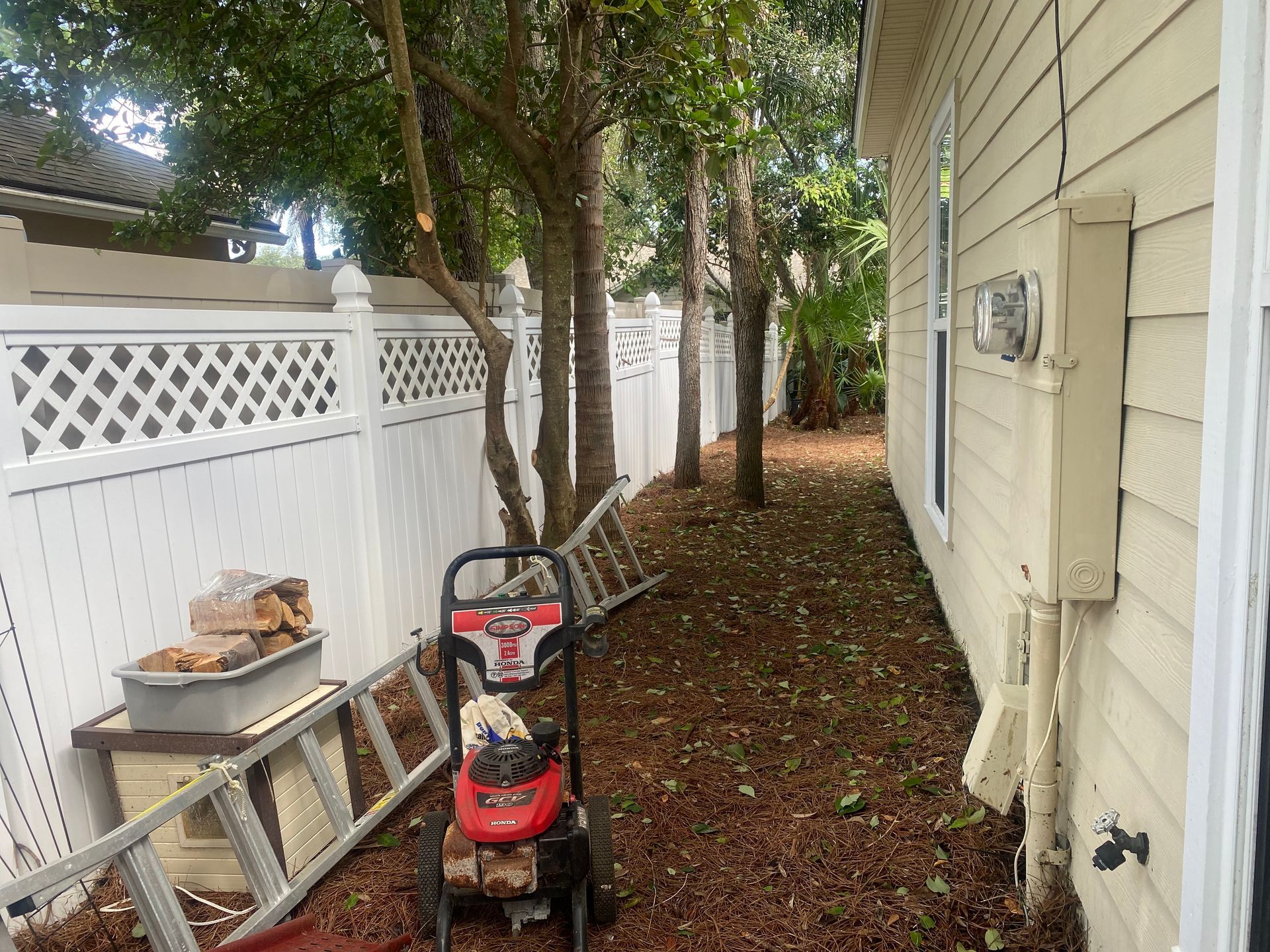 Backyard with white fence, trees, a ladder, and a power washer. House on right.