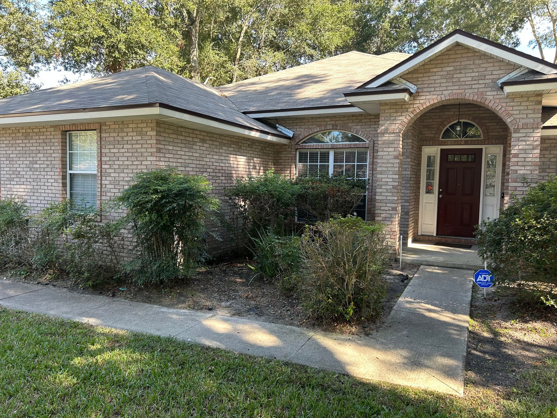 Brick house with front door, sidewalk, and overgrown bushes. Green lawn.