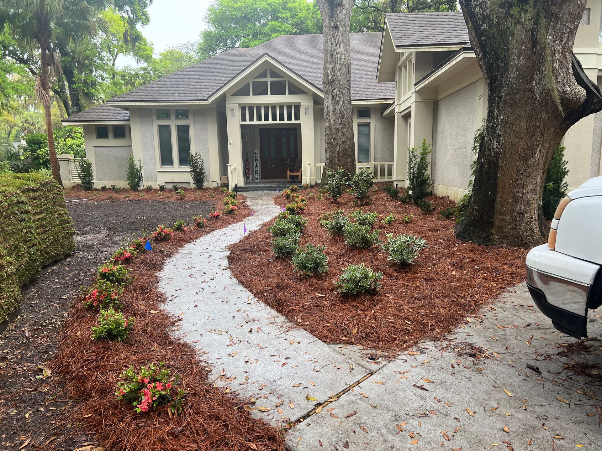 Curved white walkway to a beige house with mulched flower beds and trees in front yard