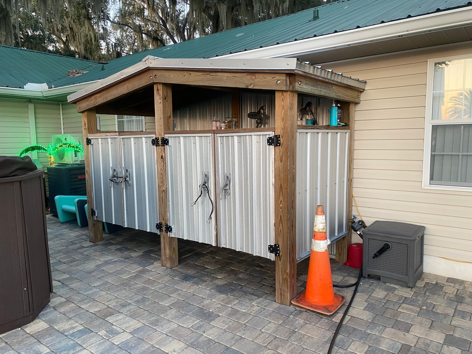 Outdoor shower with corrugated metal walls and wooden frame, on a patio. Orange traffic cone nearby.