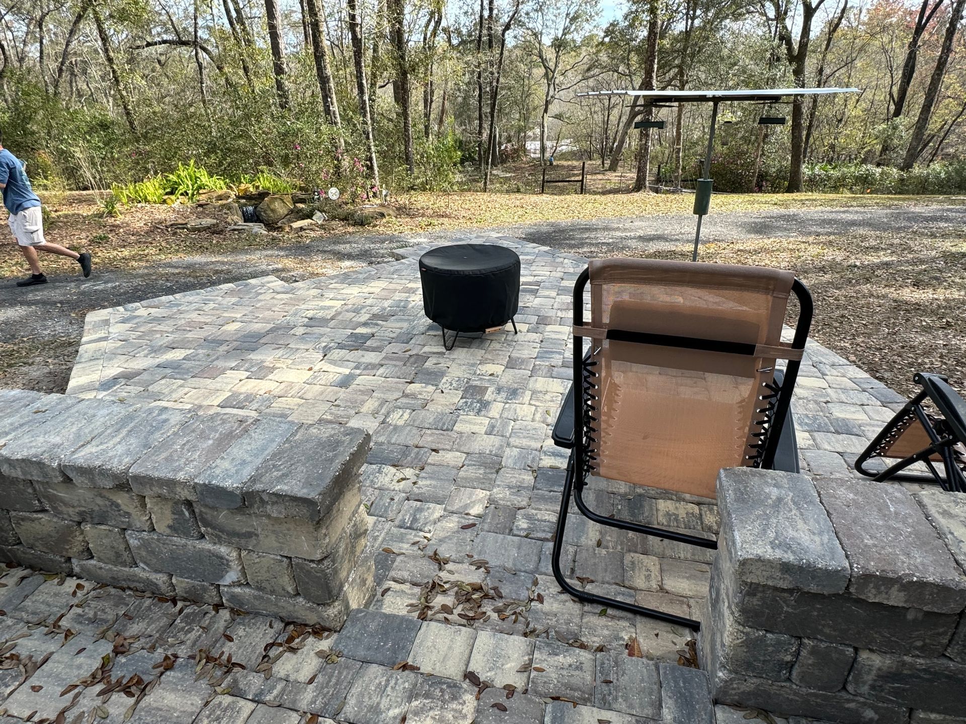 Patio with brick pavers, fire pit, chair, and a person walking in a wooded area.
