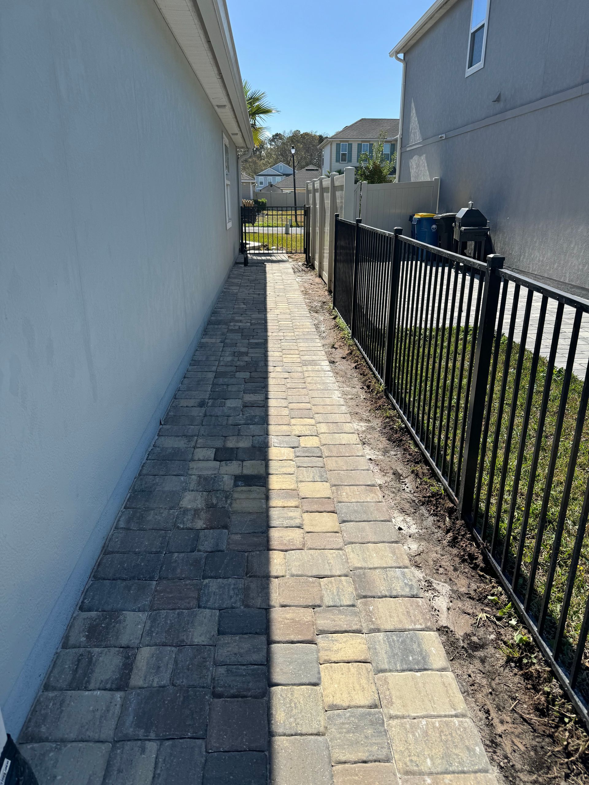 Brick pathway between a white wall and a black metal fence on a sunny day.