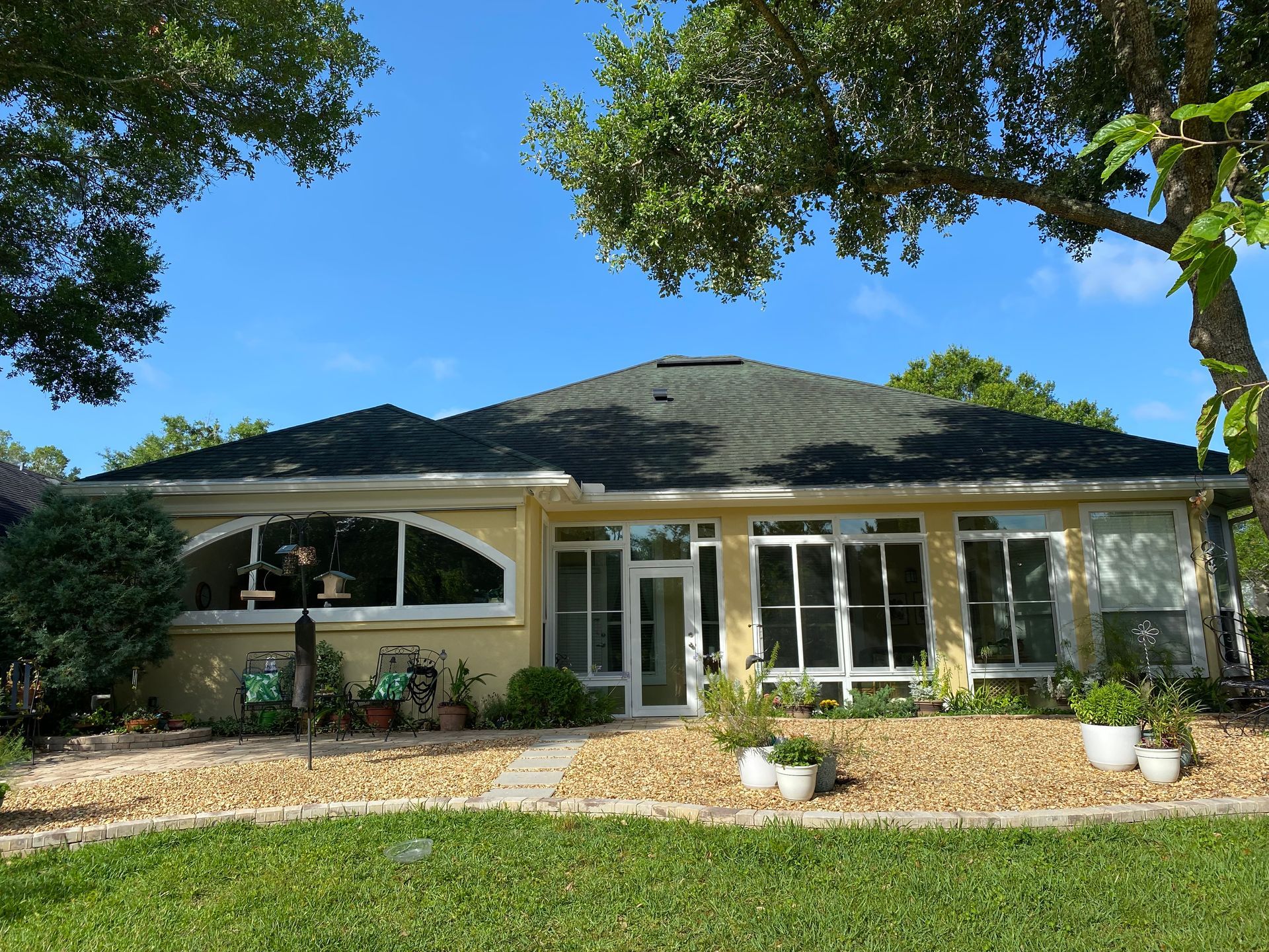 Yellow ranch house with dark roof, windows, and light landscaping under a clear sky.