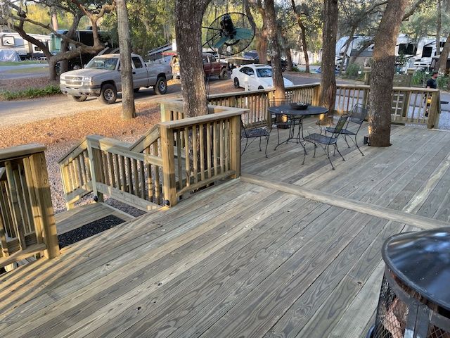 Wooden deck with railing, table and chairs, near trees and parked vehicles.