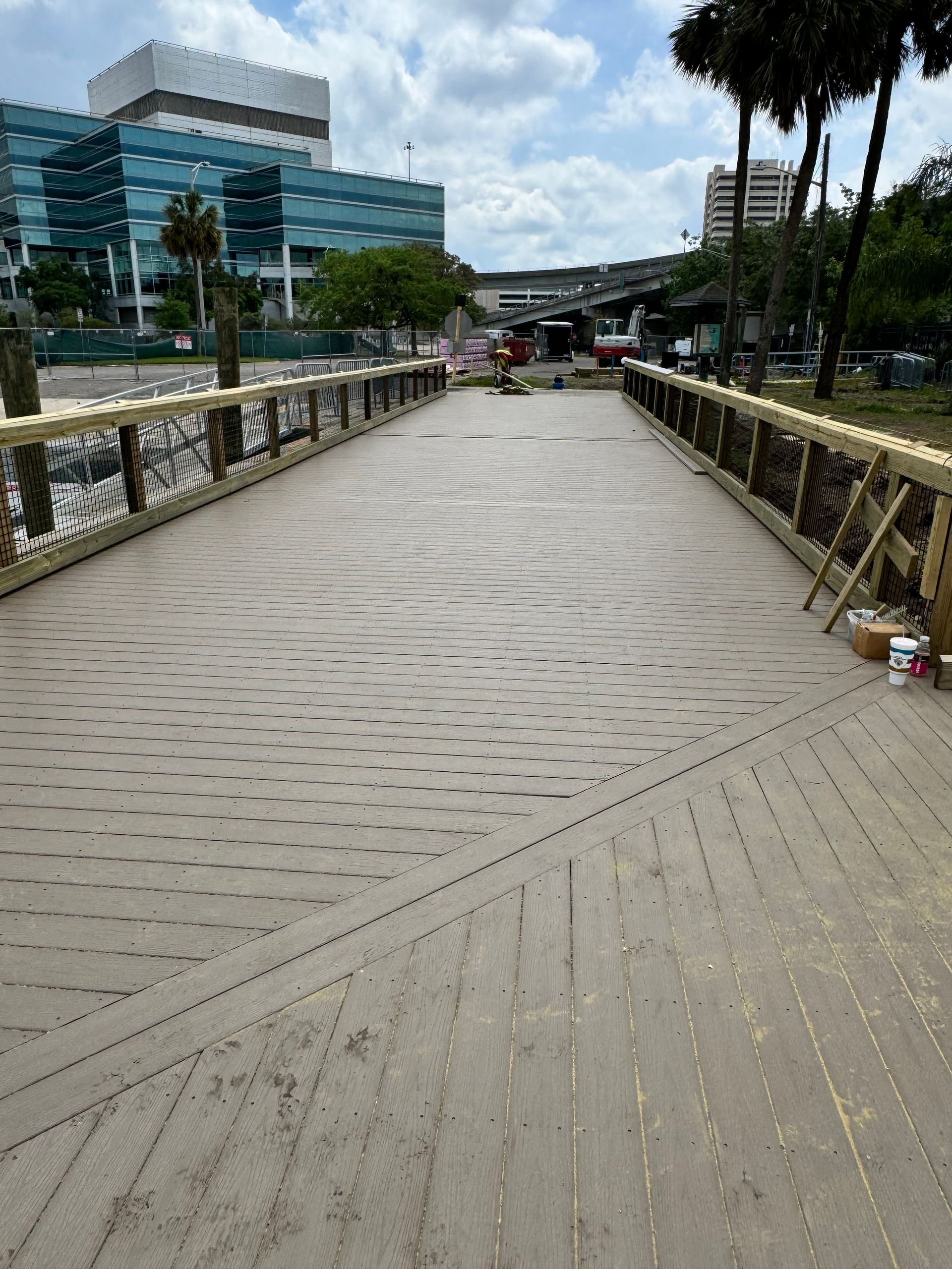 Wooden boardwalk with rope railing, leading toward buildings, palm trees, and overcast sky.