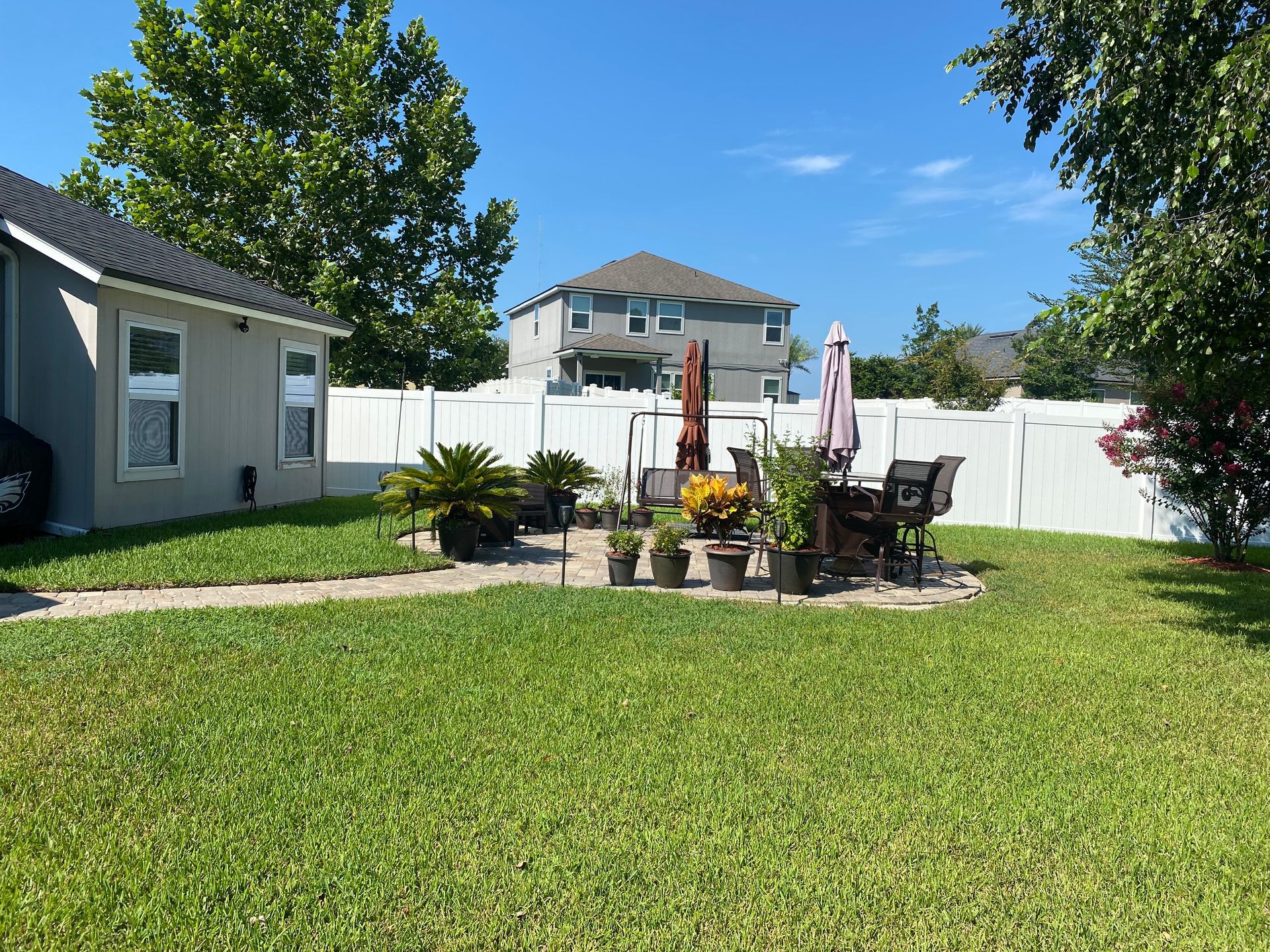 Lawn and patio with furniture, potted plants, and white fence under a blue sky.