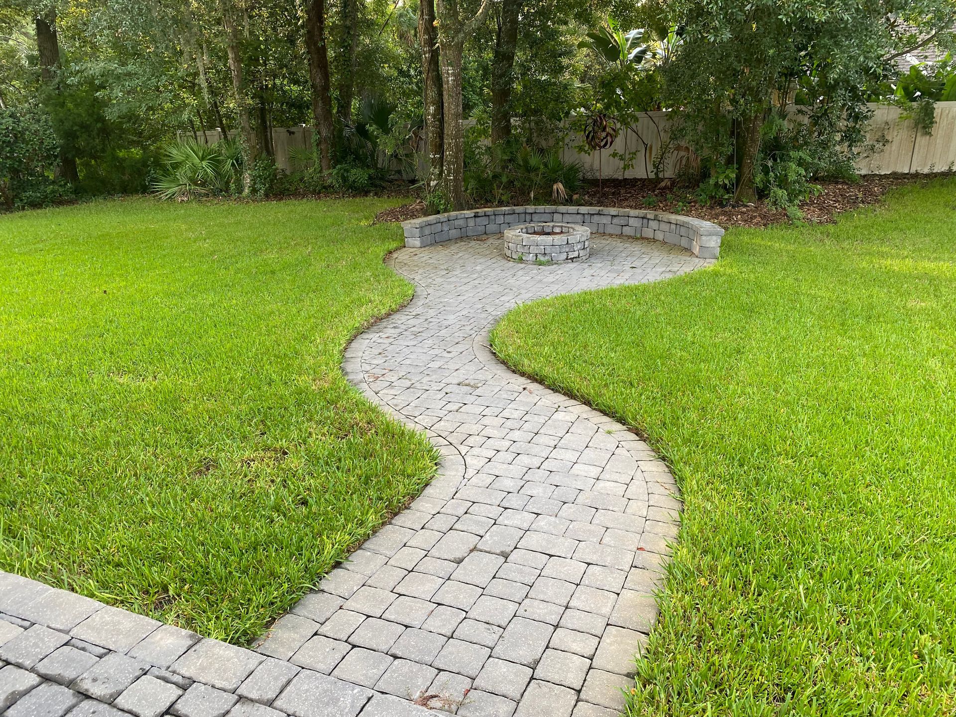 Curving brick path leads to a fire pit area in a grassy backyard, surrounded by trees.