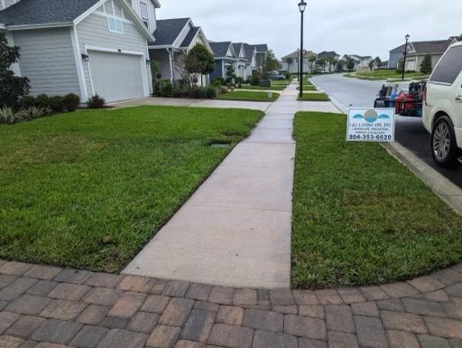 Suburban street with sidewalk and green lawns; houses line the road. Sign on lawn and vehicle parked on the side.
