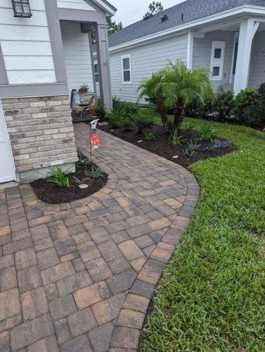 Brick pathway curves towards a house with lush landscaping and lawn.
