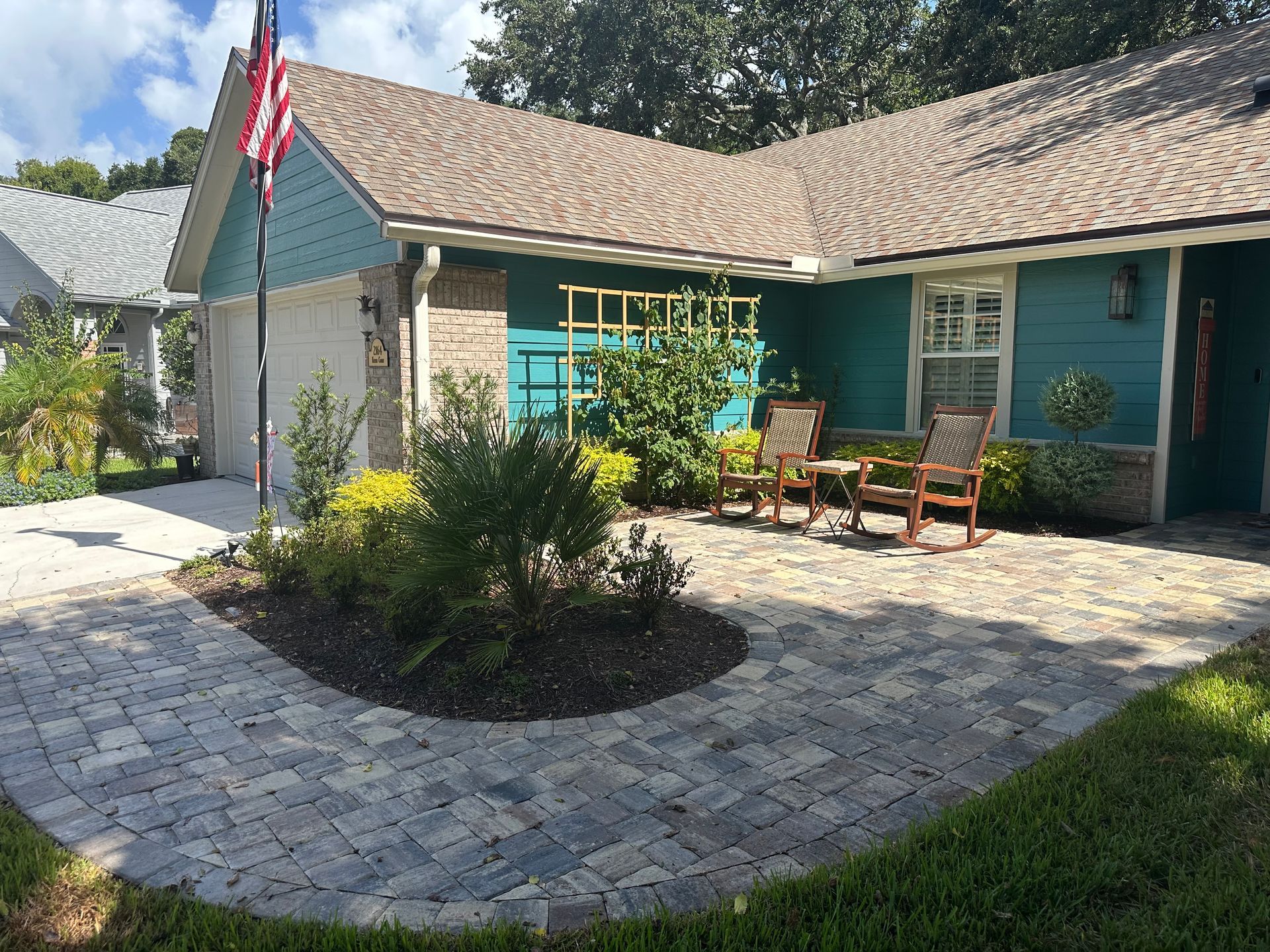 Paver patio and walkway leading to a teal house with an American flag. Two chairs on patio.