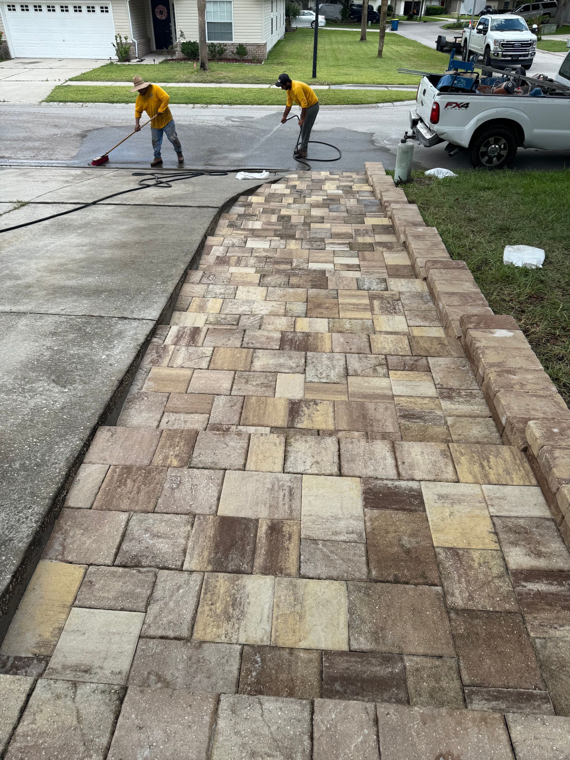 Two workers sweep a newly laid brick walkway next to a driveway.