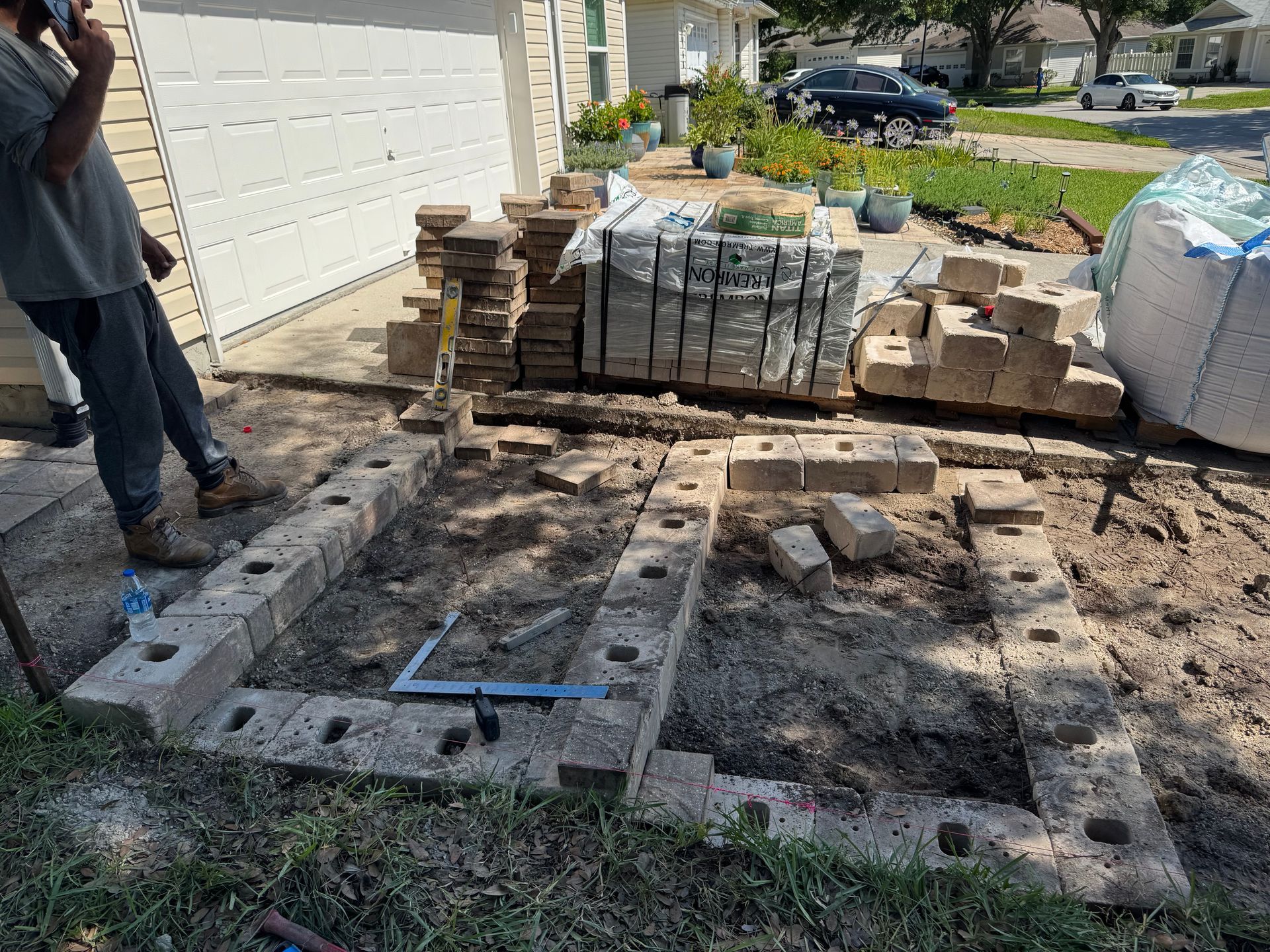 A person building a brick structure on dirt, with bricks and supplies nearby.