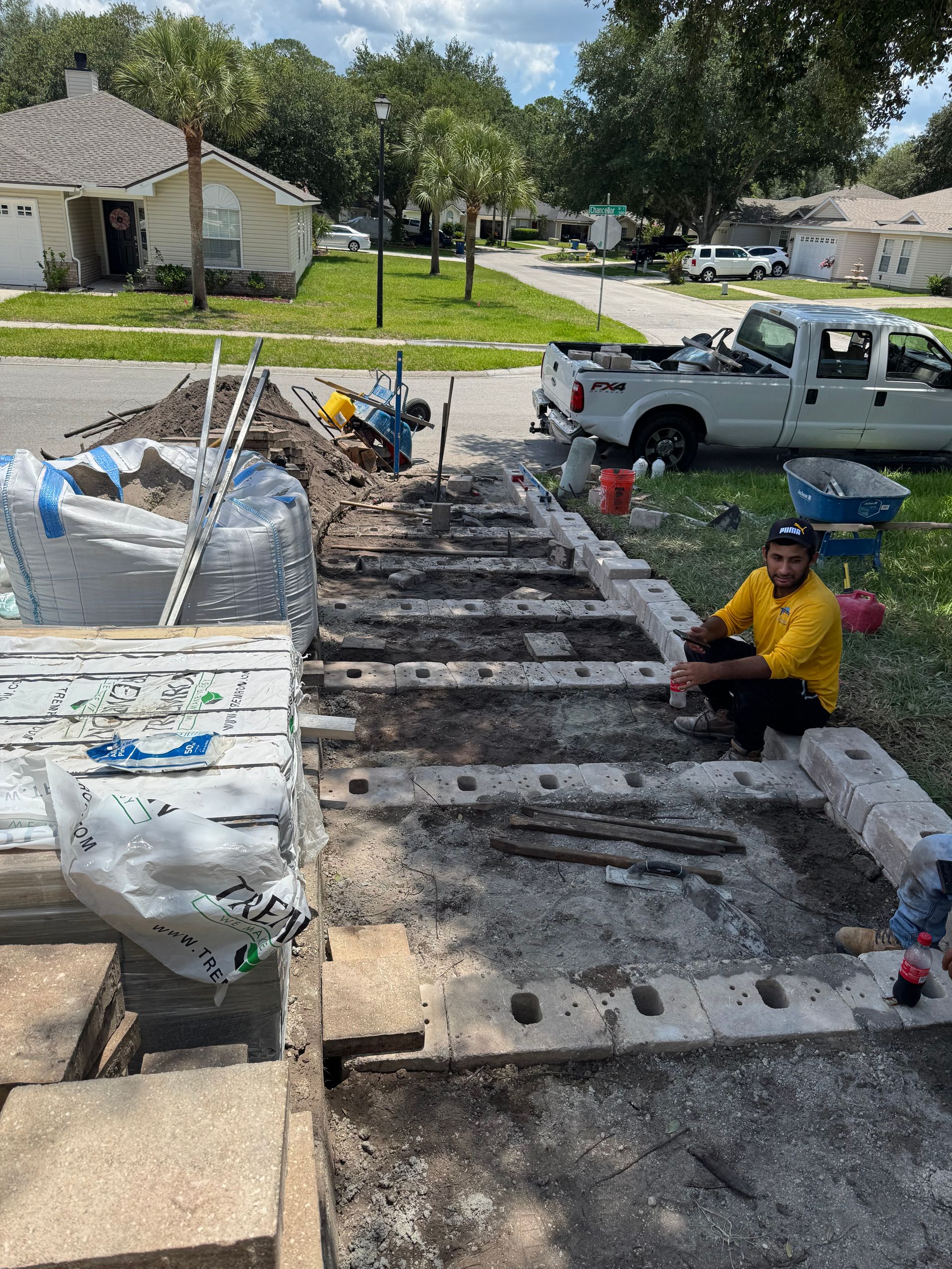 Construction worker sitting on steps; brickwork partially dismantled, materials and truck visible on sunny day.