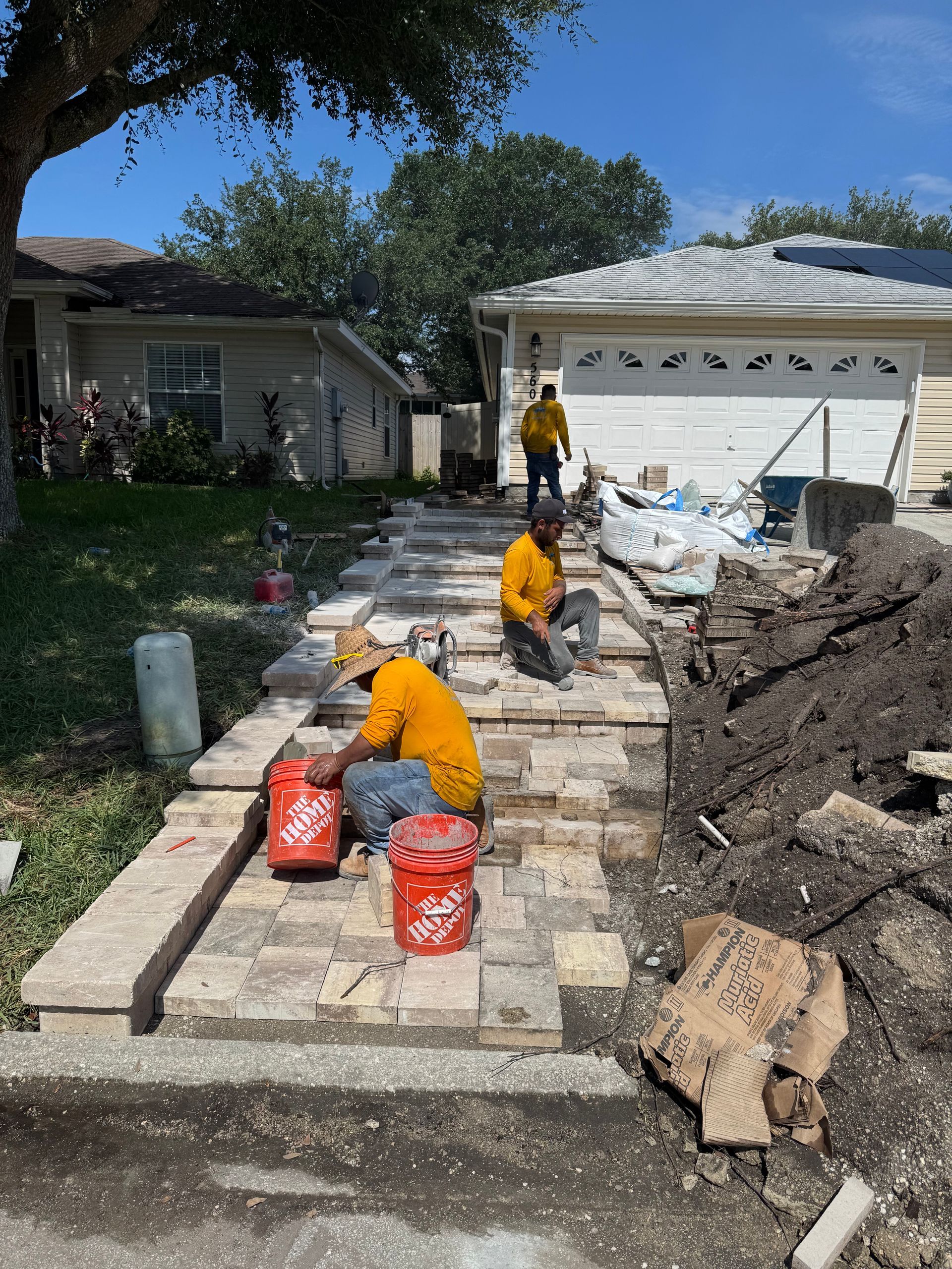 Workers laying pavers on a driveway in front of a house, with orange buckets and tools.