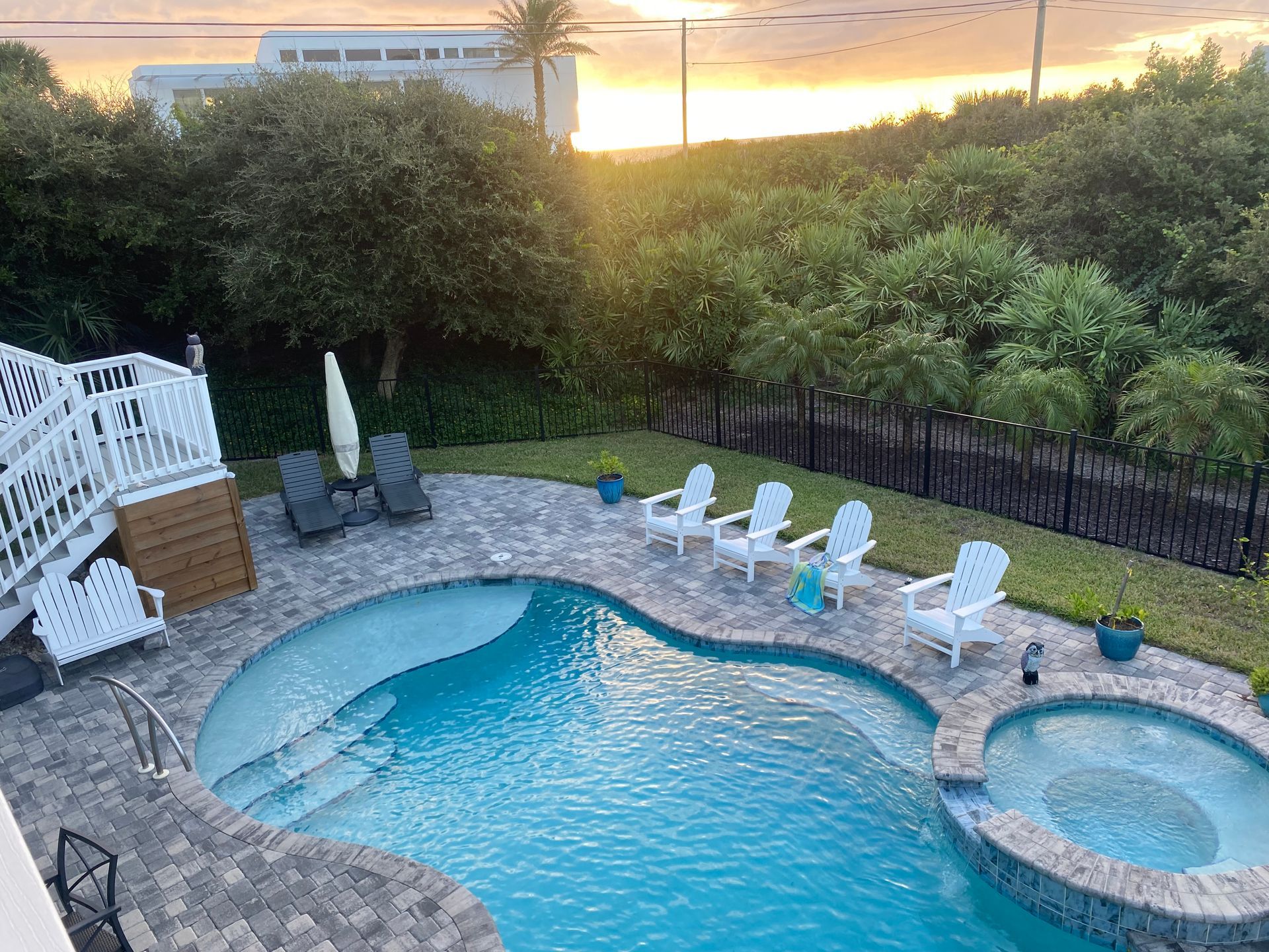 Backyard pool with white chairs, view of the ocean, sunset, and greenery.