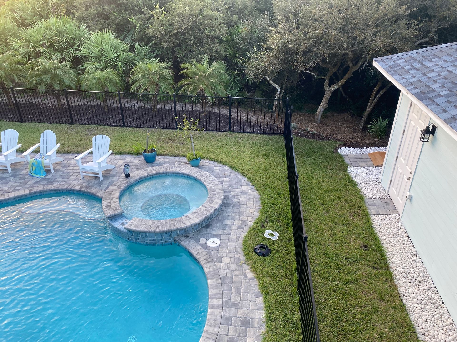 Pool and spa in a backyard with white chairs and landscaping near a fence and shed.