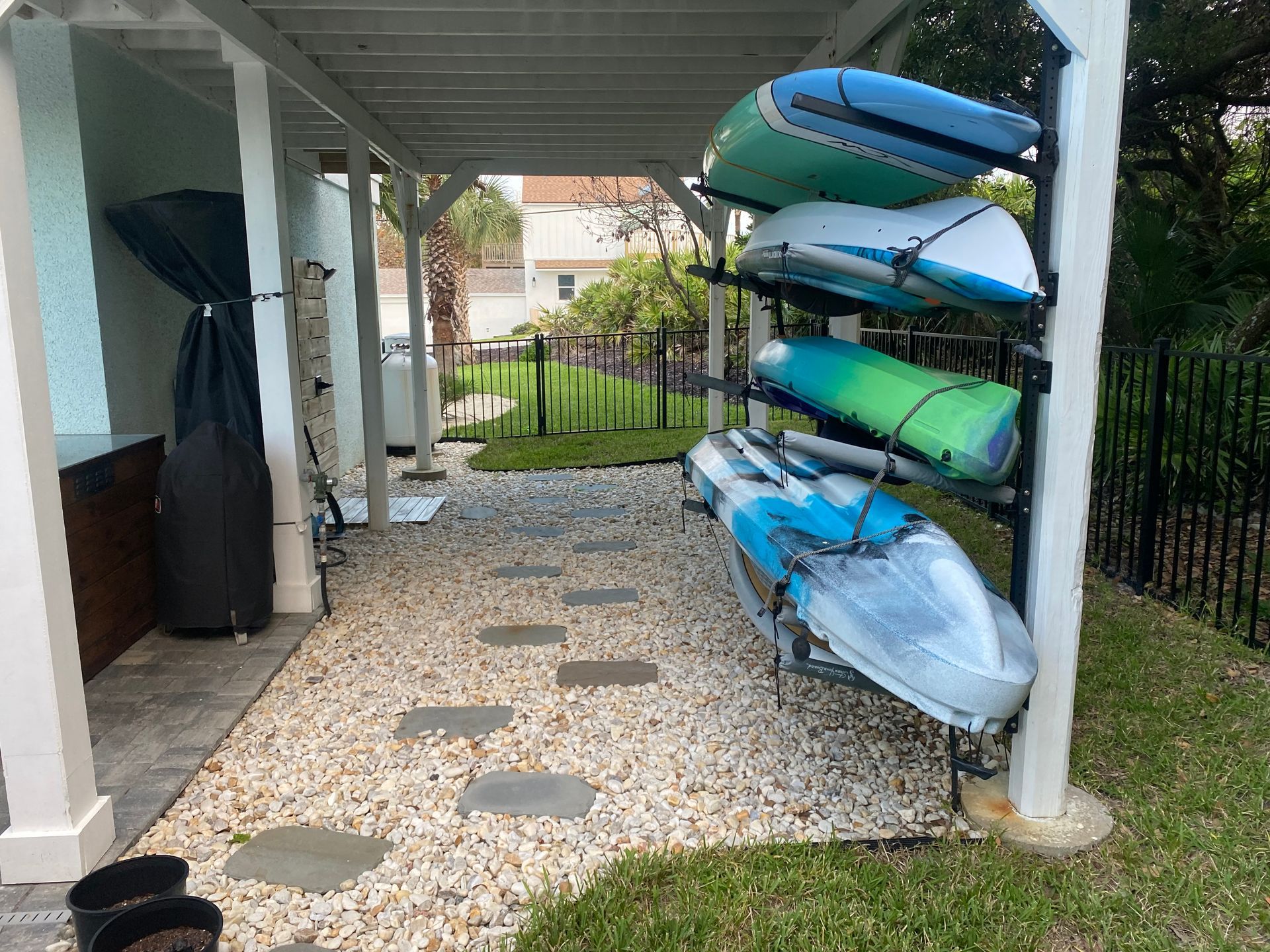 A covered outdoor storage area with a kayak rack holding four kayaks. Gravel path leads to a fence.