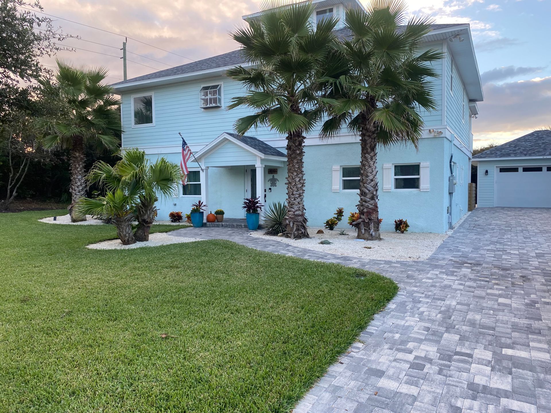 Light blue two-story house with palm trees, a brick driveway, and a well-manicured lawn.