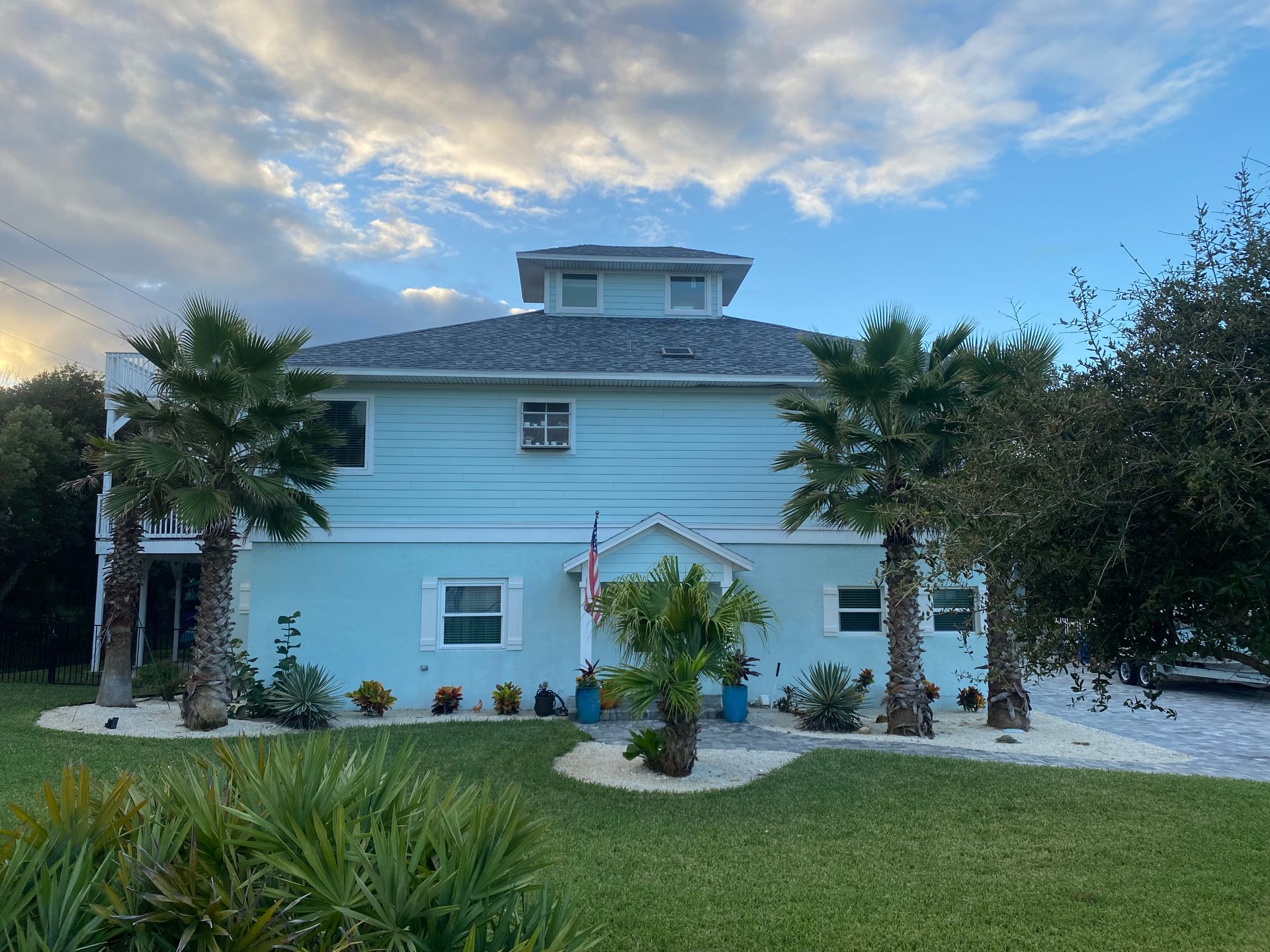 Blue two-story house with a cupola, palm trees, and green lawn under a partly cloudy sky.