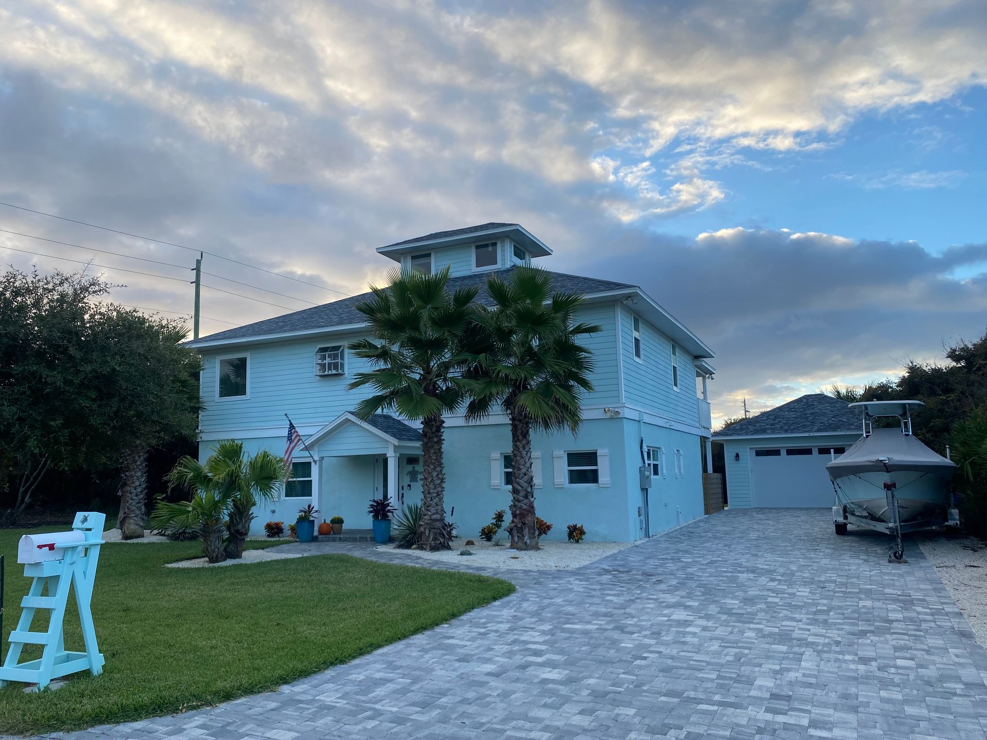 Light blue house with palm trees, driveway, and detached garage; cloudy sky.
