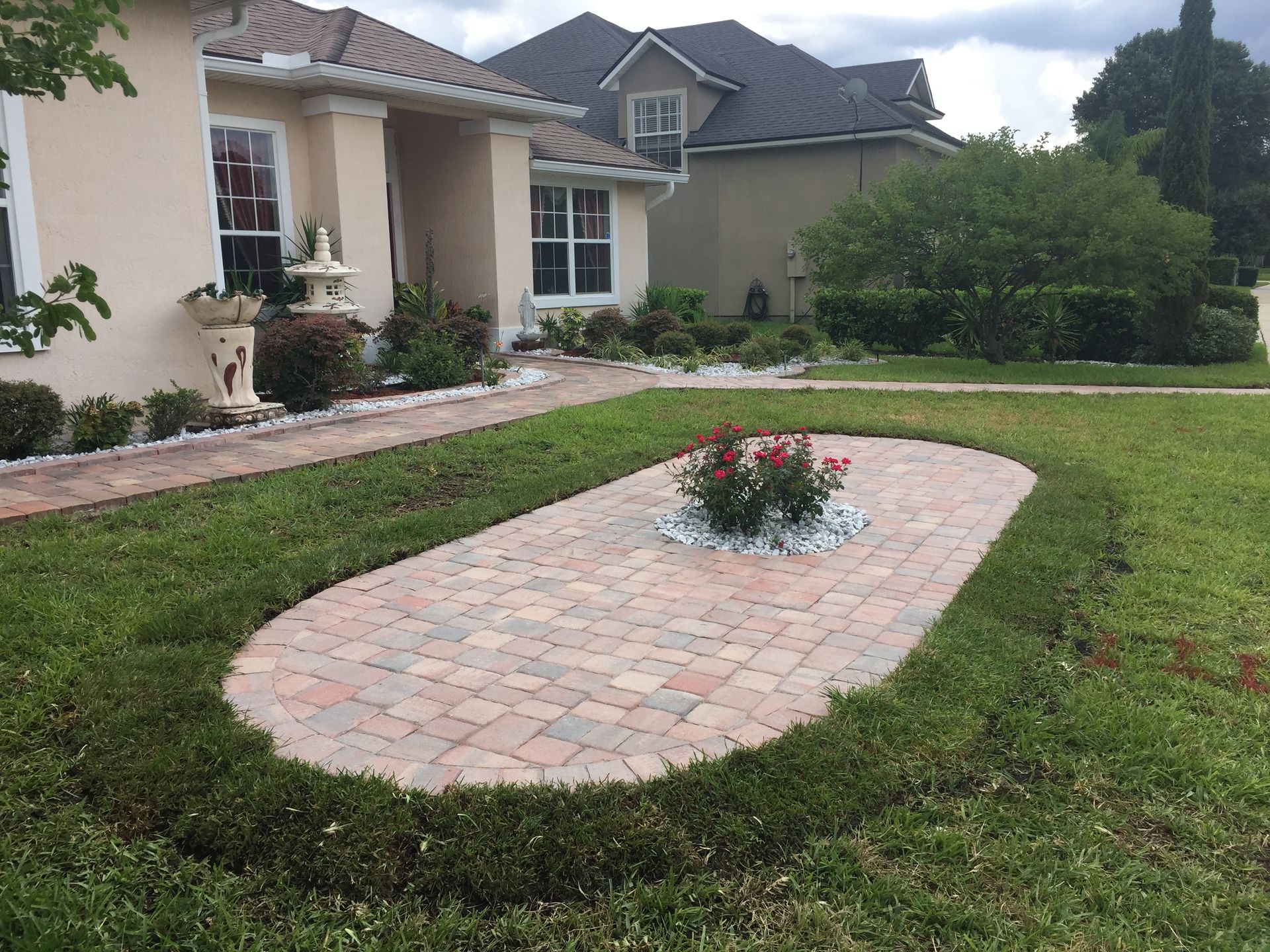 Brick walkway with rose bush, surrounded by grass and hedges, in front of a beige house.