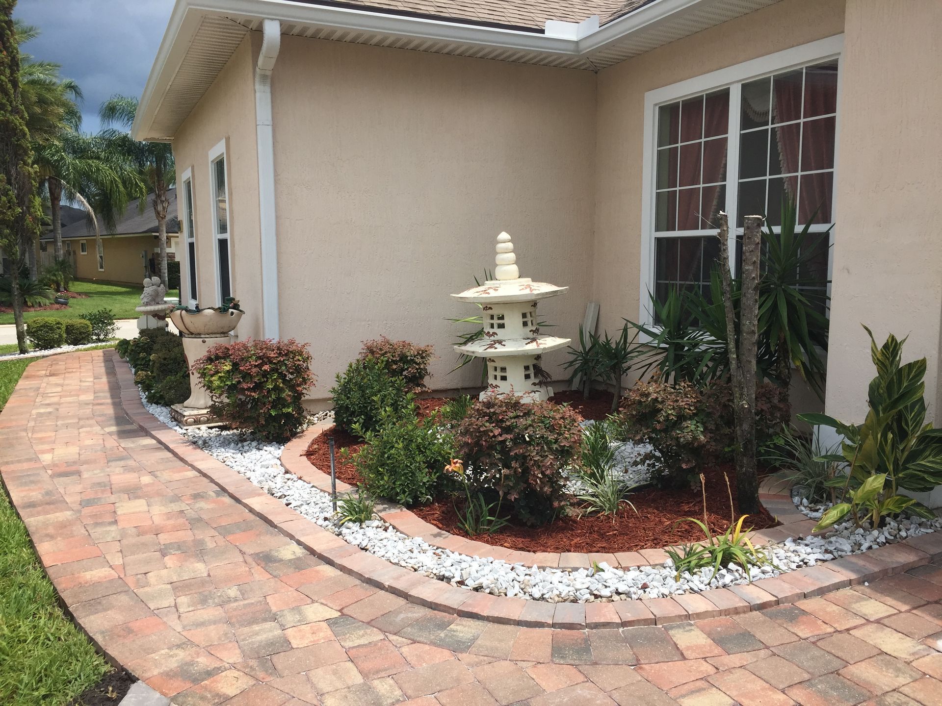 Brick walkway leading to a tan house with landscaping, a pagoda, and red and white trimmed flower beds.