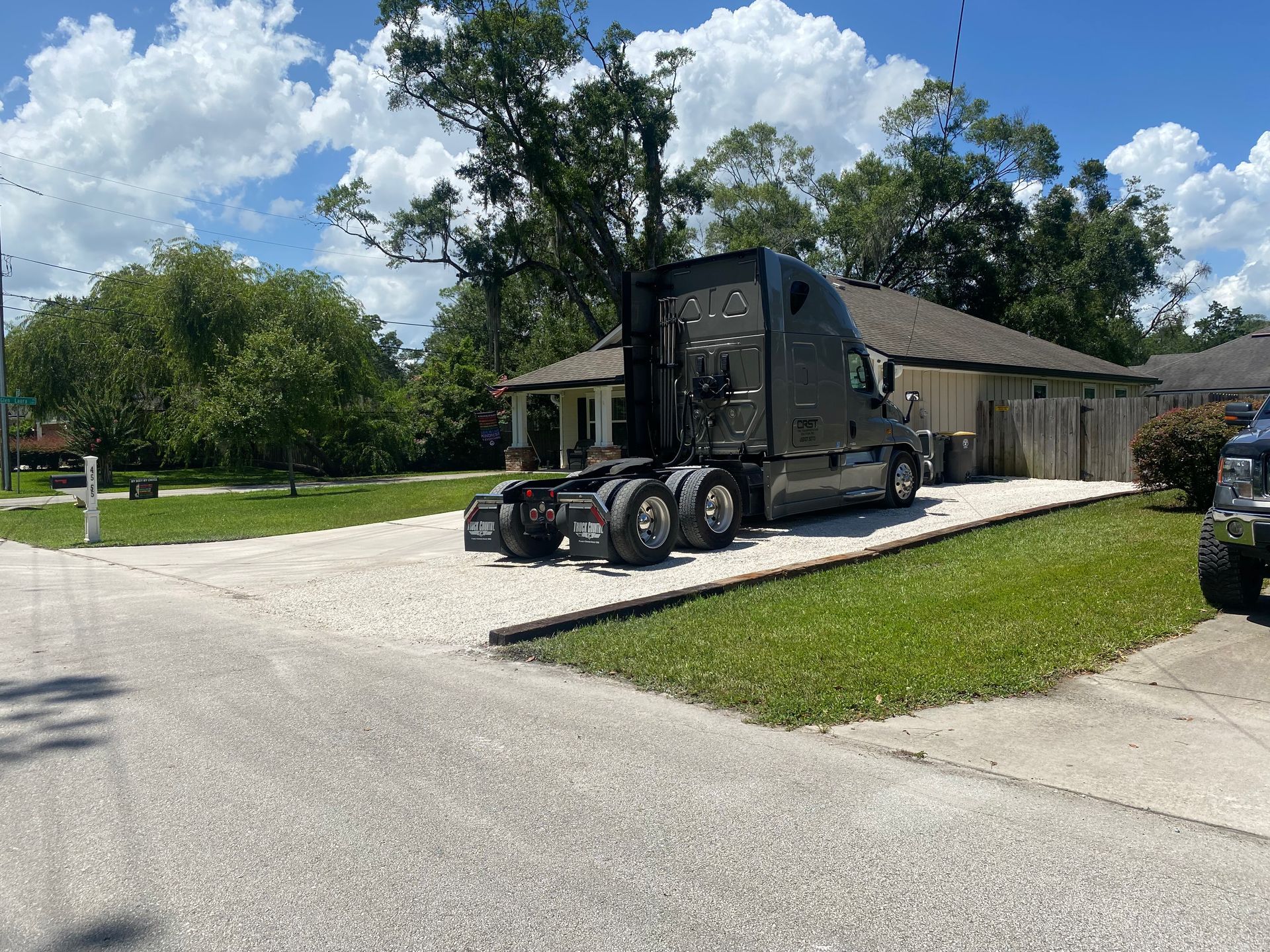 Black semi-truck parked in front of a house on a gravel driveway, under a cloudy blue sky.