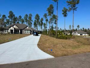 A long concrete driveway curves toward a house with gray siding. Tall pine trees are in the background.