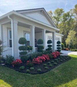 White house with porch, manicured landscaping with topiary shrubs and red flowering plants.