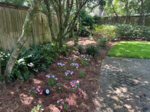 A garden bed with pink flowers, trees, and a brick pathway next to a green lawn.