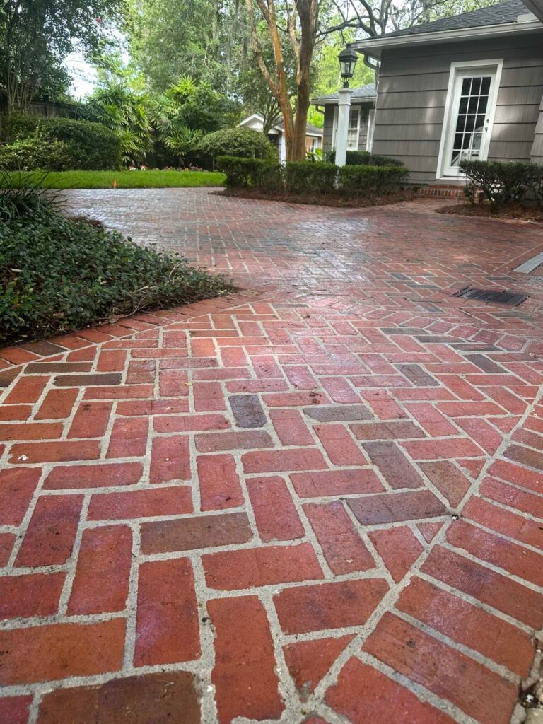 Red brick herringbone driveway with green shrubs and a grey building in background.
