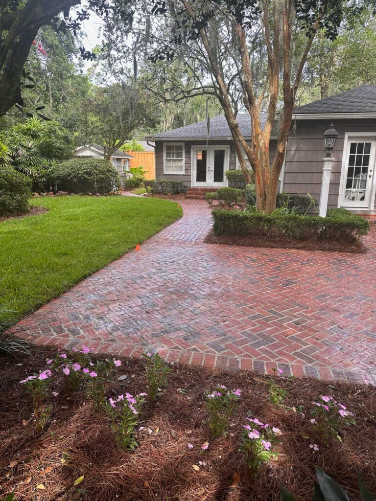 Brick walkway leading to a house with a tree in front. Green grass, bushes, and flowers.